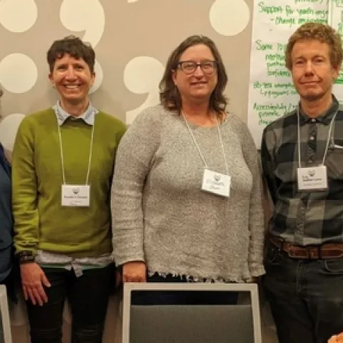 Six people wearing name tags stand in front of a wall that is partly covered in white poster paper with discussion notes written in green ink.