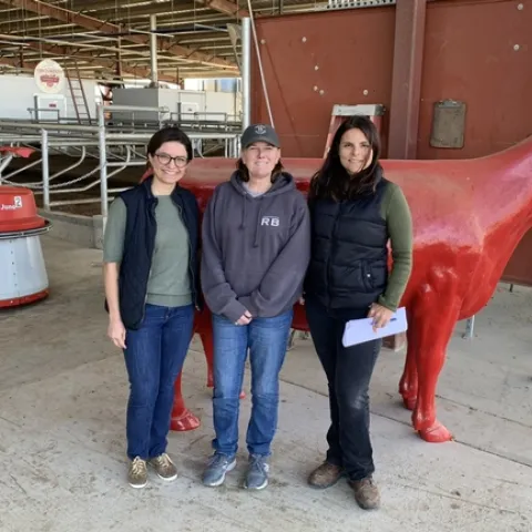 Three women stand together in a dairy barn with a feeding robot in the background