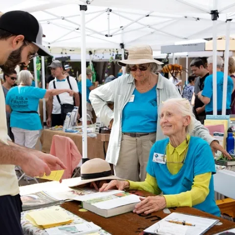 A woman is sitting down at a table, talking to a young man who is standing on the other side of the table.