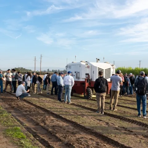 Dozens of people gather around a large machine rolling through an unplanted field and