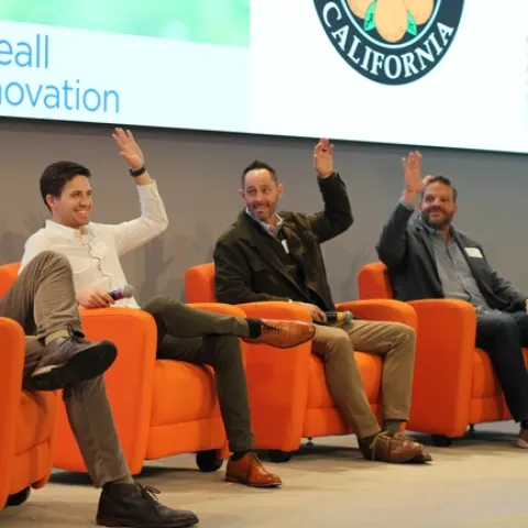 Five middle aged men sit on stage, raising their hands.