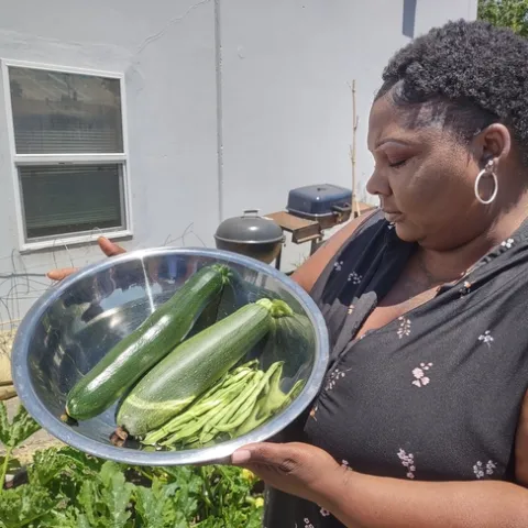 A Black woman, standing in a garden, holds a stainless steel bowl filled with zucchini and beans