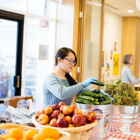 A woman wearing blue gloves sacks zucchini in a metal bucket on a table of baskets of oranges, apples, broccoli and carrots.