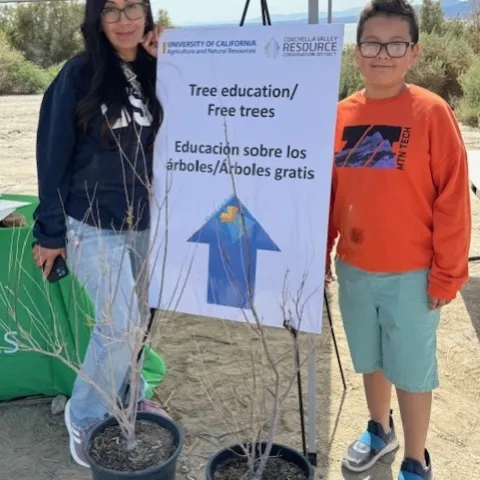 A woman and boy stand beside a sapling and sign that reads: Tree education/free trees. Educacion sobre los arboles/arboles gratis.