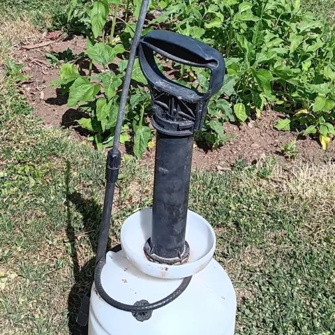 A hand-pump sprayer with the handle extended and spray nozzle resting against the tank, surrounded by lawn.