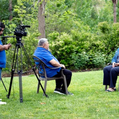 A man standing behind a camera records another man interviewing a woman while sitting down.