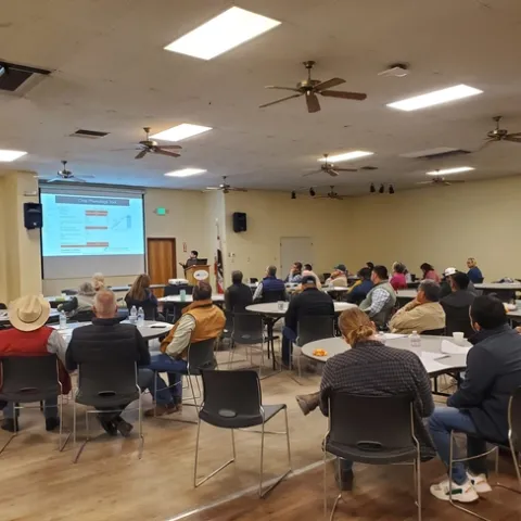 A room full of mostly men sitting around round tables look at a PowerPoint slide as Tapan Pathak describes a crop phenology tool.