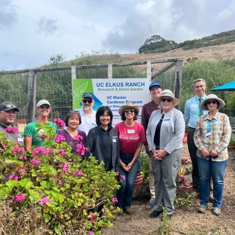 Ten people stand next to a flowering hot pink geranium bush. Hanging on the fence behind them is a sign that reads, "UC Elkus Ranch research & demo garden. UC Master Gardener Program. San Mateo & San Francisco counties