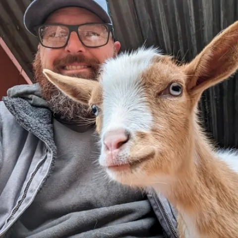 A man with a hat, glasses and a beard next to a brown and white goat.