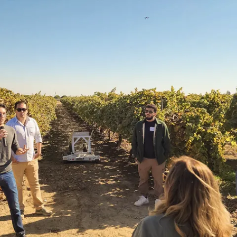 A man standing in a vineyard speaks into a mic as people listen. The Rover robot rolls between rows of grapevines.