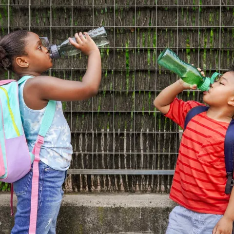 Two children wearing school backpacks drinking water from their reusable water bottles.