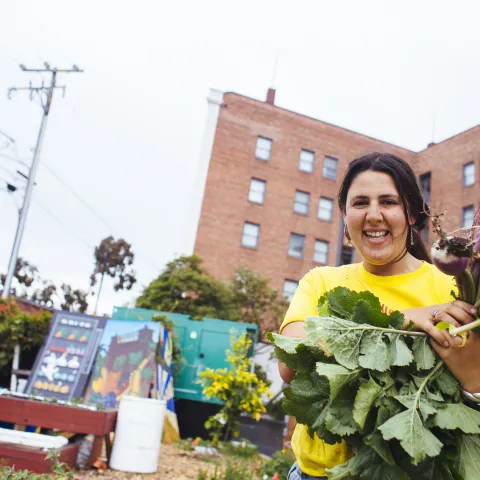 A farmer presents a crop from an urban plot in front of a brick building