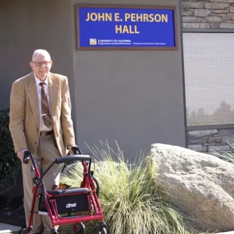 John Pehrson, standing behind a walker, smiles under the John E. Pehrson Hall sign on the building named in his honor.