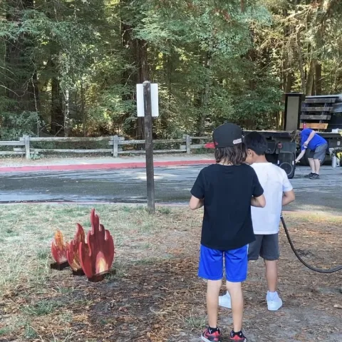 Two boys, one aiming a water hose at wooden cutout of flames