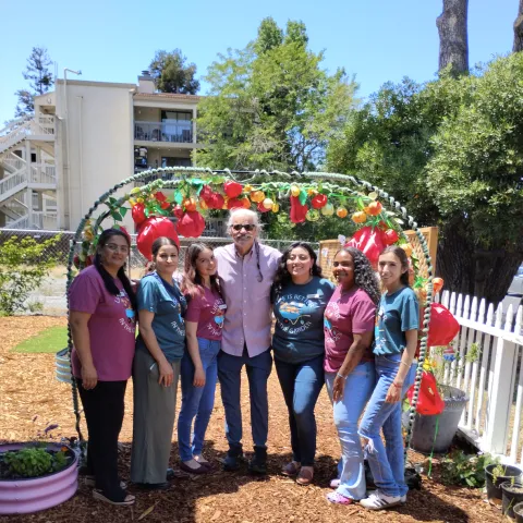 Santos stands beneath an arch decorated with red, yellow and green vegetable ornaments. Three women stand on both sdies of him