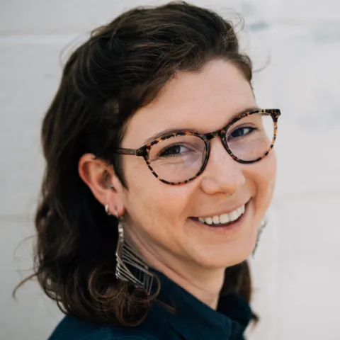 A brown-haired white femme wearing glasses and earrings smiles at the camera.