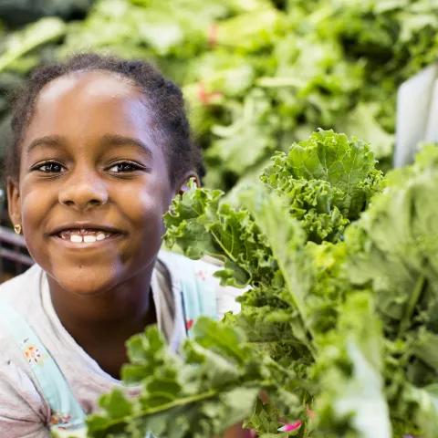 Girl smiles and holds up some leafy greens at a market