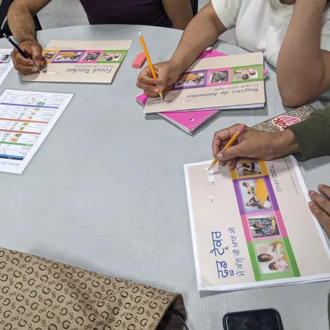 Three people with their hands writing at the same table on worksheets that depict three different languages 