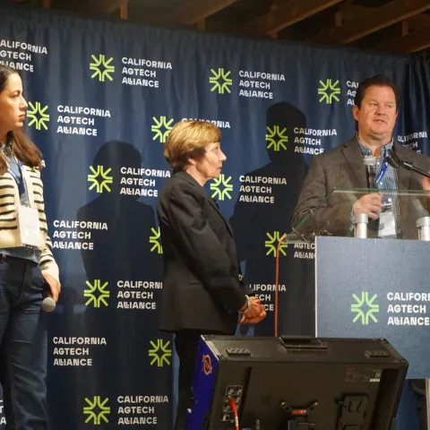 Gabe Youtsey speaks at a podium while Karen Aceves and Karen Ross look on at AgTech Alliance press conference