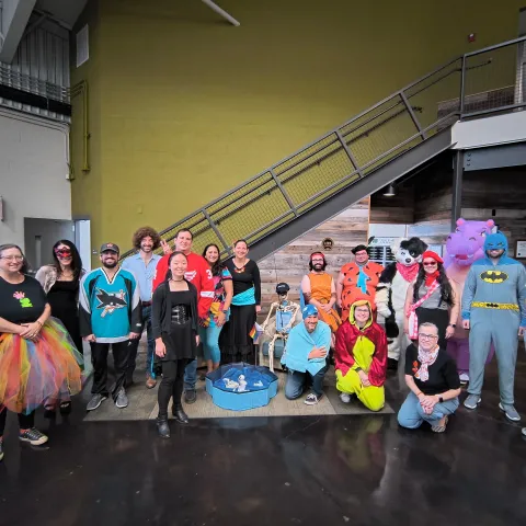 19 people dressed in costumes pose with a skeleton next to stairs in ANR building foyer