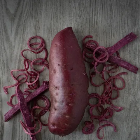 A whole dark purple-skinned sweetpotato flanked by pieces of purple sweetpotato flesh cut into curls