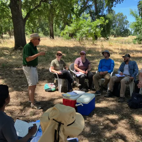 Man in polo and shorts and hat at left speaks with a group of students under shade trees during a farm tour