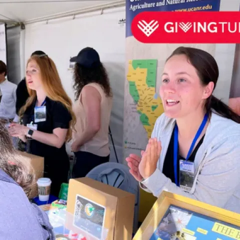 A woman speaks to someone visiting an event booth. Text: GivingTuesday