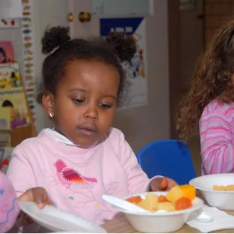 A young girl sitting at a table with other young children grabs a bowl of fruit.