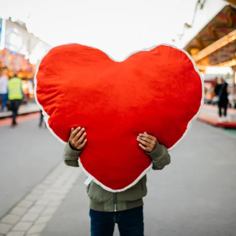 person holding a large stuffed heart covering their face on a busy street