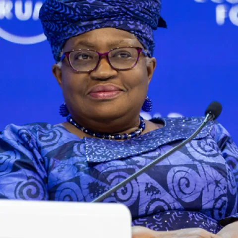 Ngozi Okonjo-Iweala sitting at the podium of the world economic forum in 2021. She is wearing a blu dress with swirl patterns and a blue head covering with a smile on her face. 