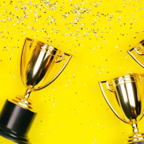 Gold trophy cups scattered diagonally across a bright yellow background, with silver confetti sprinkled around them.