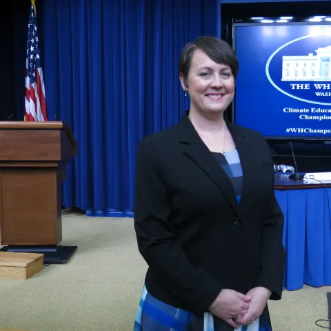 Woman wearing a black blazer smiles as she stands in a White House meeting room with a podium and table in the background
