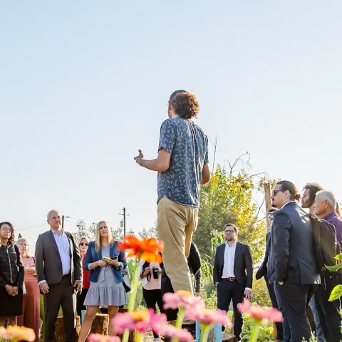 A speaker addresses a group of visitors gathered among rows of corn, sunflowers, and zinnias on a small farm in California's Central Valley