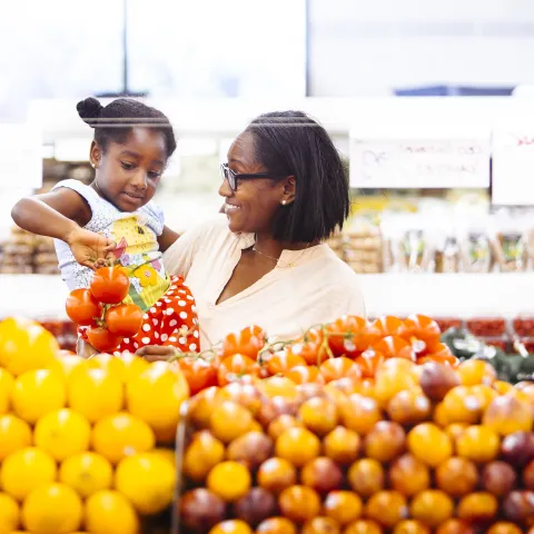 A mother holds her young daughter in her arms while selecting apples in the produce section of a grocery store.