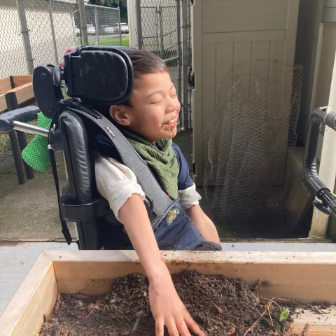 Young boy seated and strapped into a wheelchair seated adjacent to an elevated garden box, touching dirt with his left hand and smiling with joy