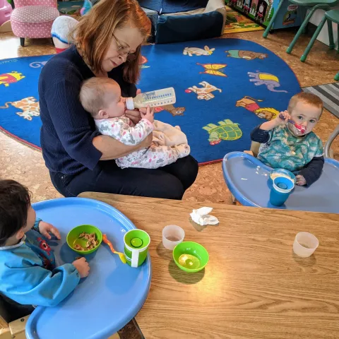 A childcare provider holds an infant in her arms while bottle feeding her. Two toddlers sit next to her in high-chairs eating snacks.