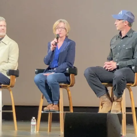 A woman seated on a stool onstage speaks into a mic as men on either side of her watch her speak
