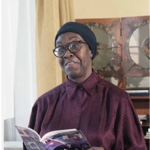Gwendolyn Brooks sitting in a room holding a book wearing a red shirt, glasses and a blue headscarf