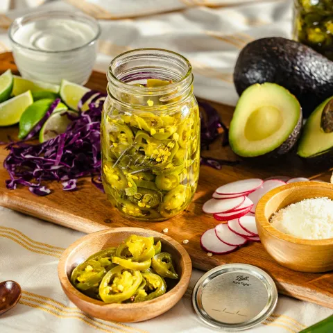 a jar of sliced jalapenos sits on a cutting board surrounded by shredded red cabbage, lime wedges, bowl of salt, sliced radishes, halved avocados, and tortillas