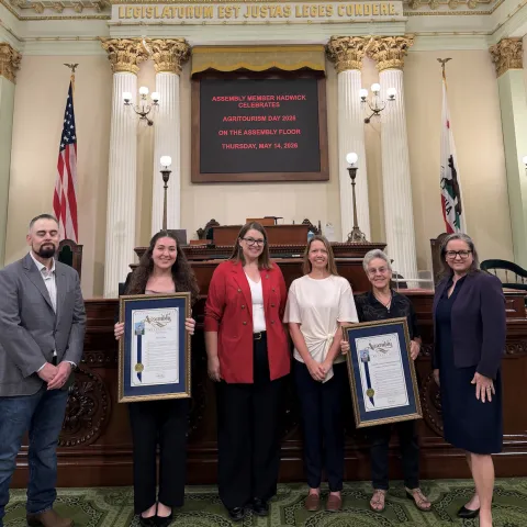 Six people stand in front of U.S. flag in the Assembly chamber. Two of the women are holding framed resolutions.