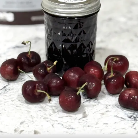 scattered cherries in front of a glass jar of cherry jam