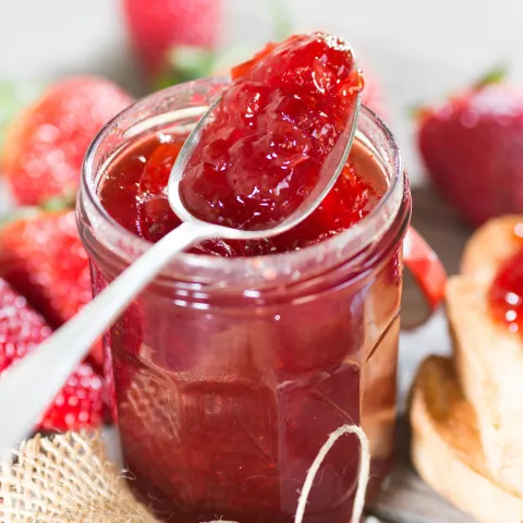 Jar of strawberry jam with spoon scooping some out, and strawberries lying around the jar. 