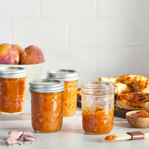 4 glass jars of zesty peach bbq sauce on a countertop with a bowl of peaches in the background, next to a plate of chicken and basting brush