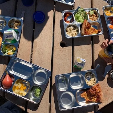 Reusable stainless steel trays filled with school lunch food set atop an outdoor table.