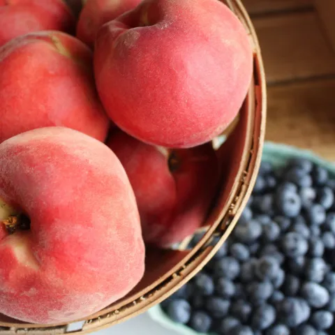 a bowl of several pink peaches next to a bowl of blueberries