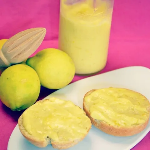 3 lemons next to a jar of frozen lemon curd and lemon juicer with two lemon cookies on a plate with a pink background