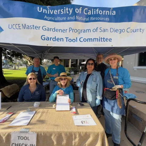 Seven people smile from under a blue canopy and white sign that reads "University of California Agriculture and Natural Resources, UCCE Master Gardener Program of San Diego County, Garden Tool Committee." The table in front of them is covered by brown burlap and has papers and a clipboard. One person is holding garden tools.