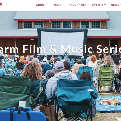 A large crowd sits in chairs facing a large outdoor movie screen set up in front of a barn-like building.