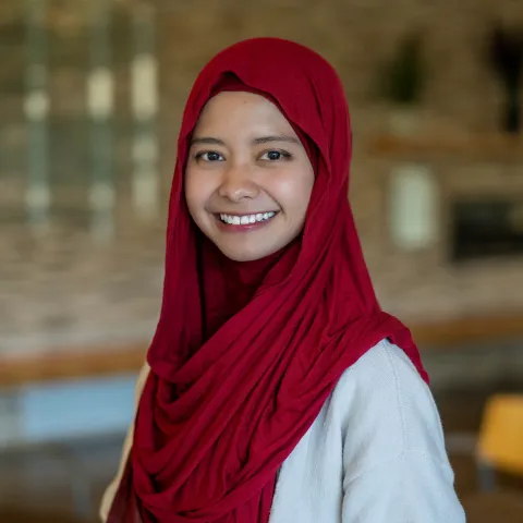 Professional headshot of a smiling woman wearing a deep red hijab and white sweater.