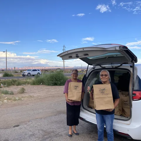 Navajo business owners standing beside a car with a paper grocery bag.